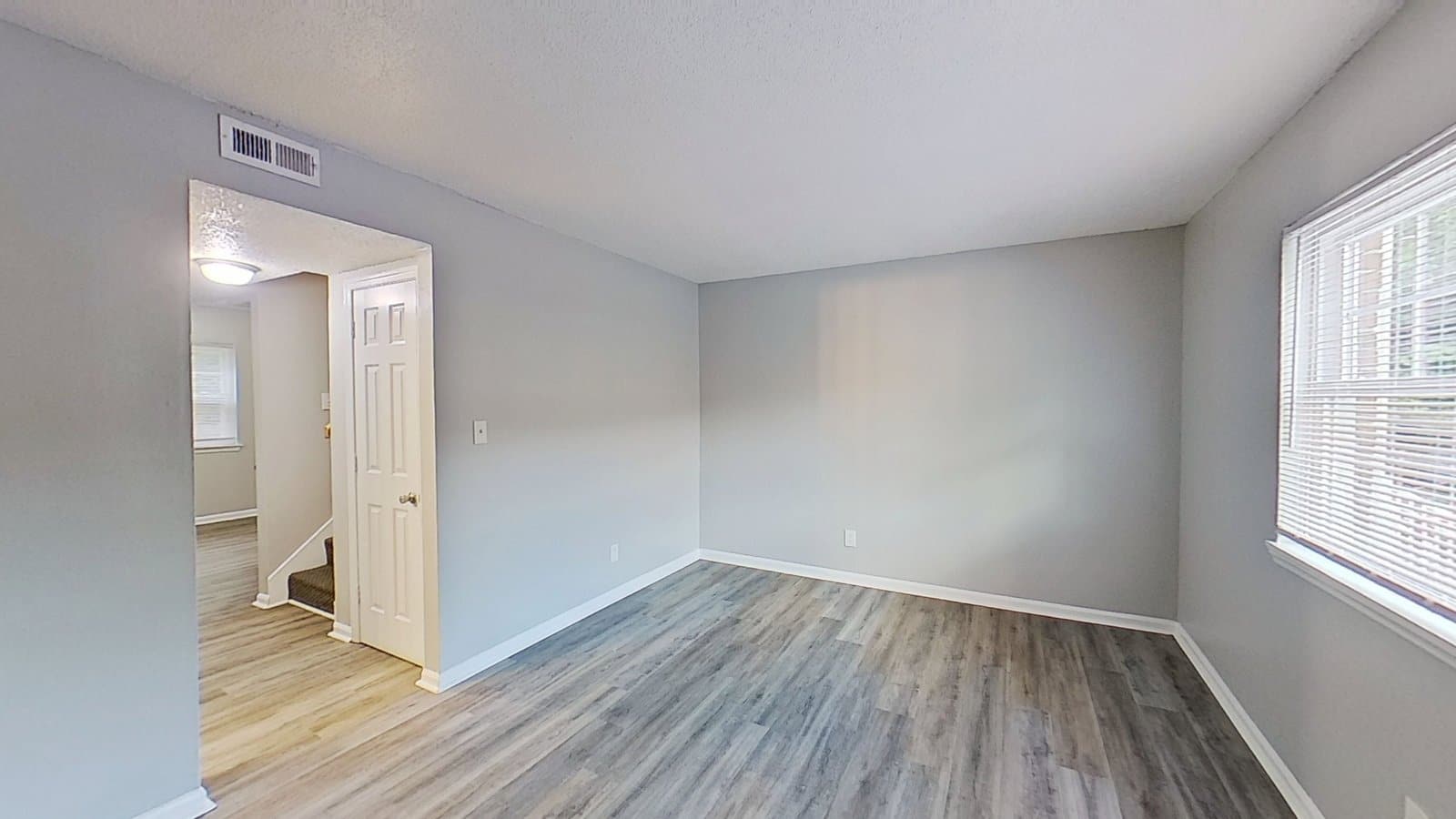 A living area inside a Harbor Pointe apartment: vinyl plank flooring, a window with blinds, and a doorway through to the stairs